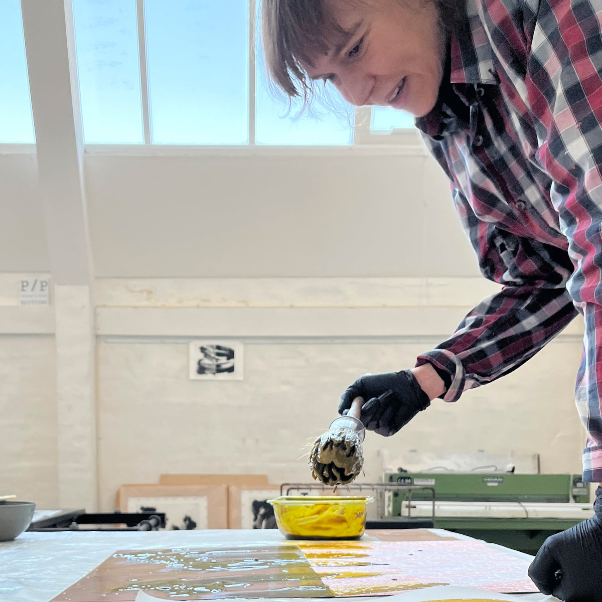 Artist Agathe de Bailliencourt leaning over a workspace at at the Printer's Proof intaglio print studio while holding a thick paint brush to paint acid onto a copper plate.