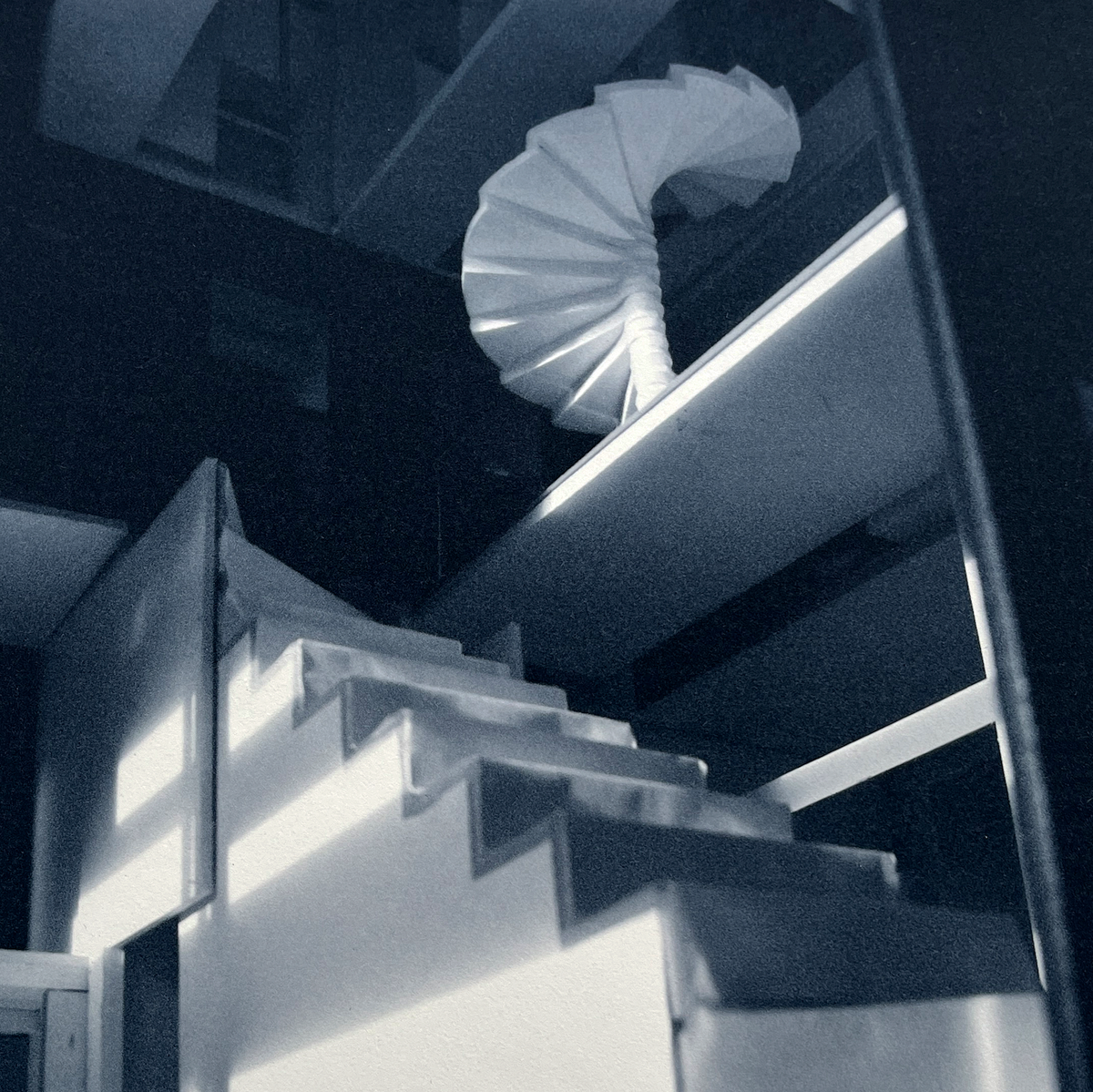 A detail of a monochrome photogravure by Anette Harboe Flensburg, depicting an empty, quiet room in rich shades of blue with a winding staircase.