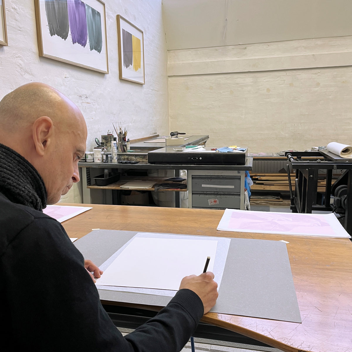 Arturo Herrera as he leans over a Printer's Proof's studio work table, signing a contemporary "Portal" print with a pencil.