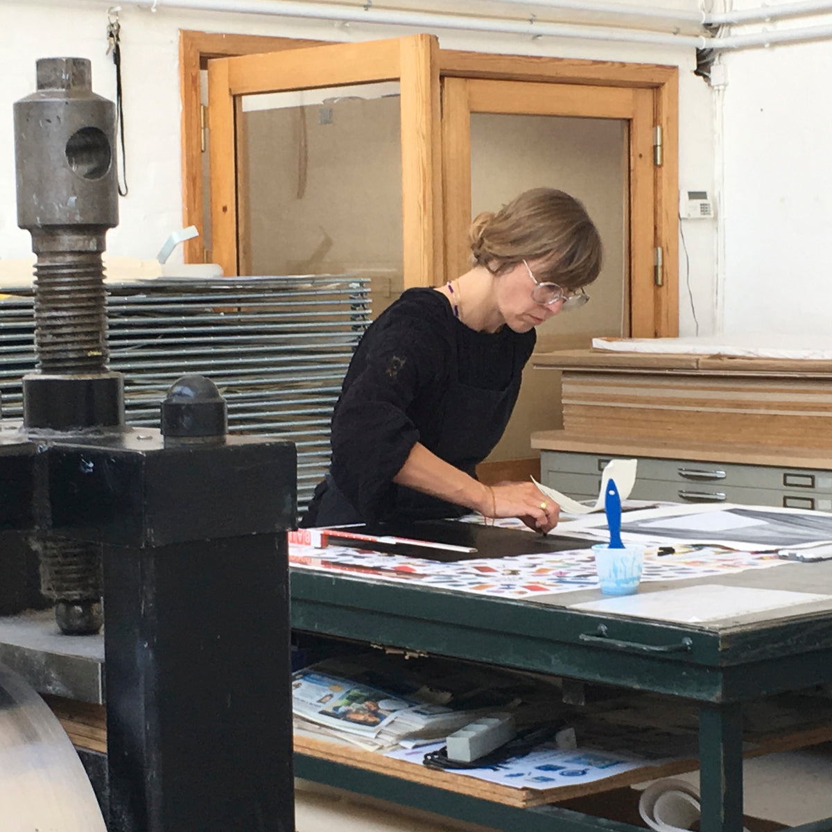 Artist Julie Sass leaning over a workspace table at Printer's Proof printmaking studio while using lithographic crayons to draw directly onto an aquatinted copper plate.