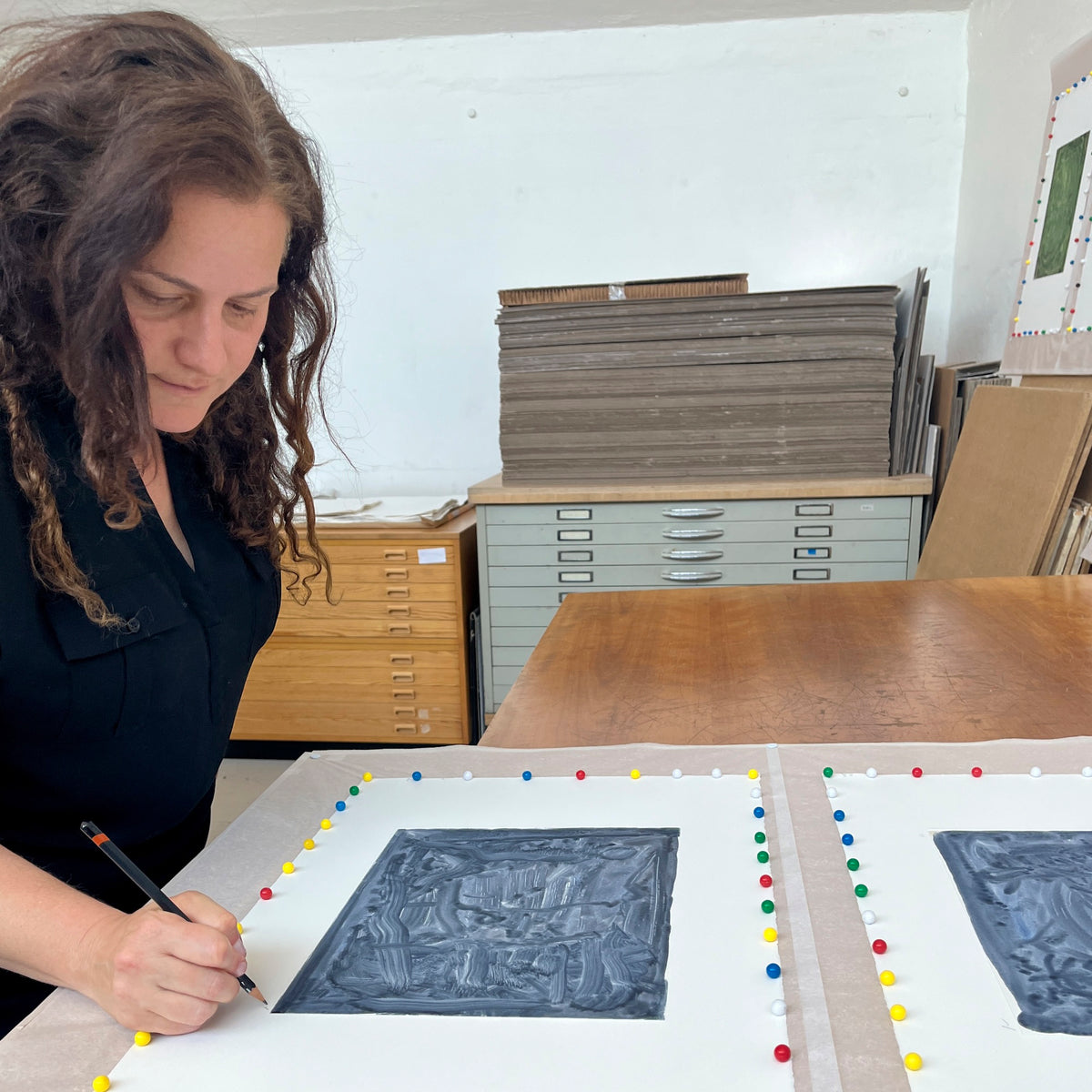Liat Yossifor leans over a Printer's Proof studio table to sign the margin of an abstract monochrome monotype.
